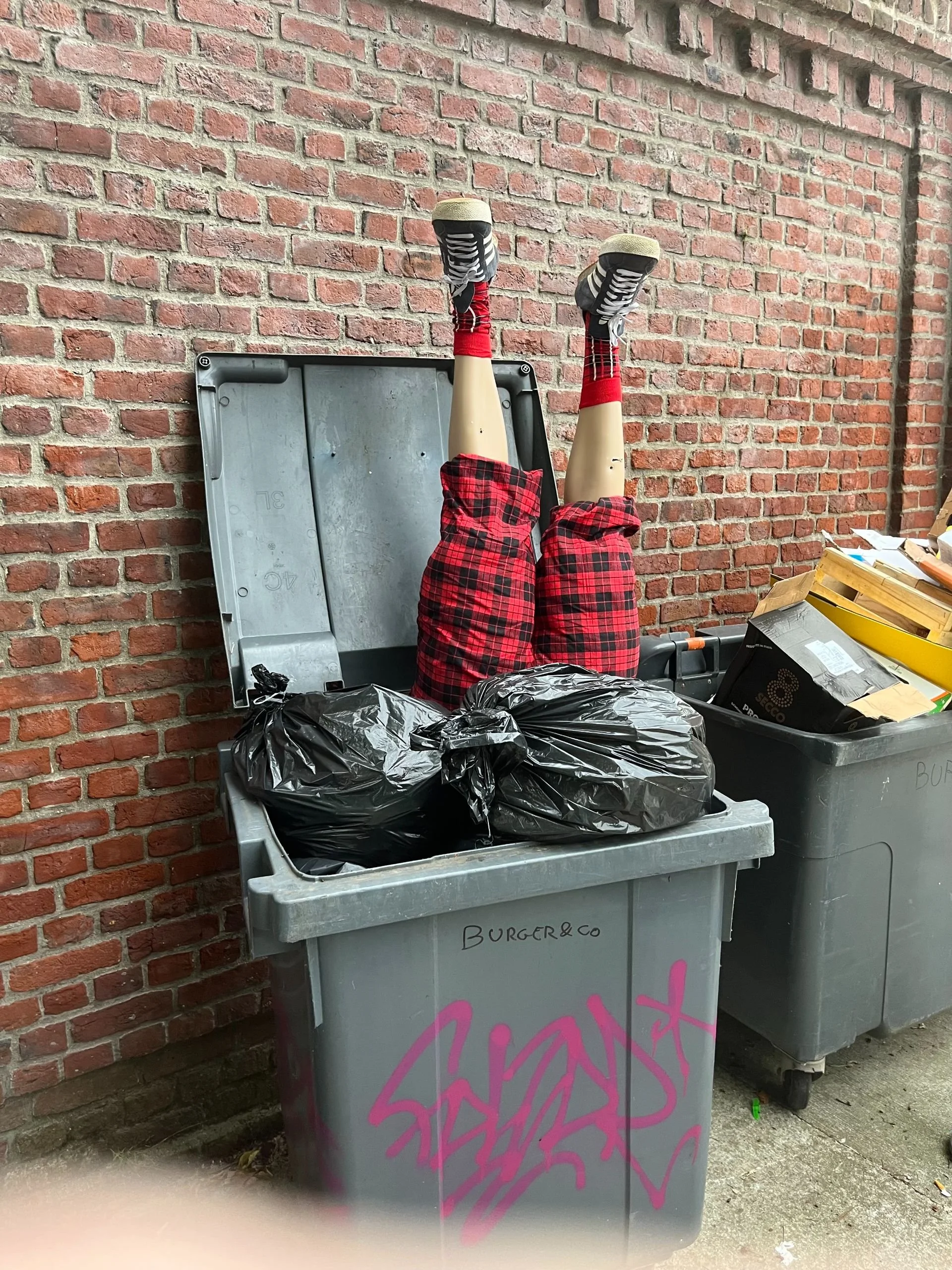 A brick wall with two large trash bins, one with black garbage bags and a pair of mannequins' legs in plaid shorts, socks, and sneakers sticking out of the bins.