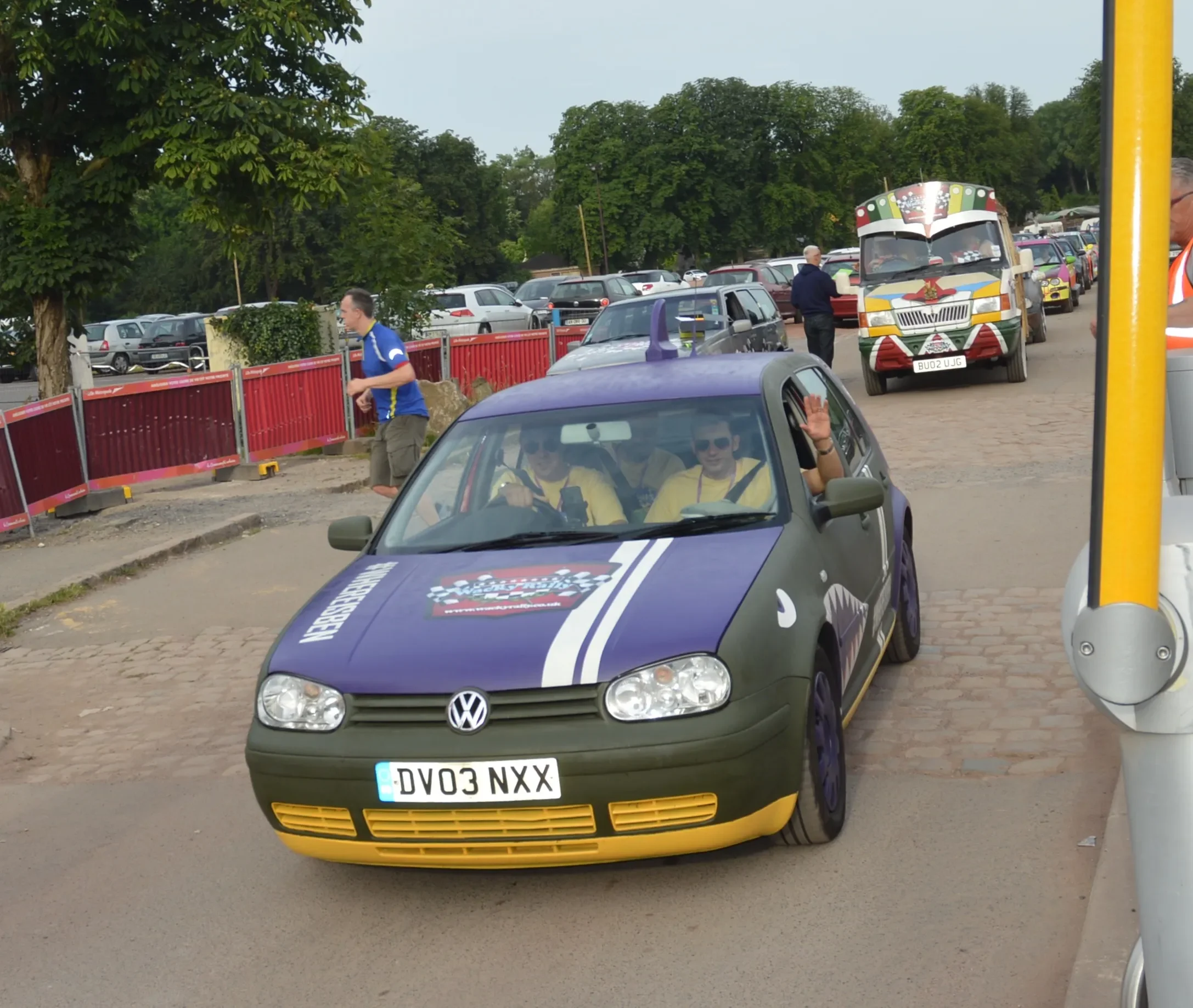 A small Volkswagen car, painted in purple and gray with yellow accents, decorated with racing stickers, and driven by people wearing yellow shirts, including one person waving.