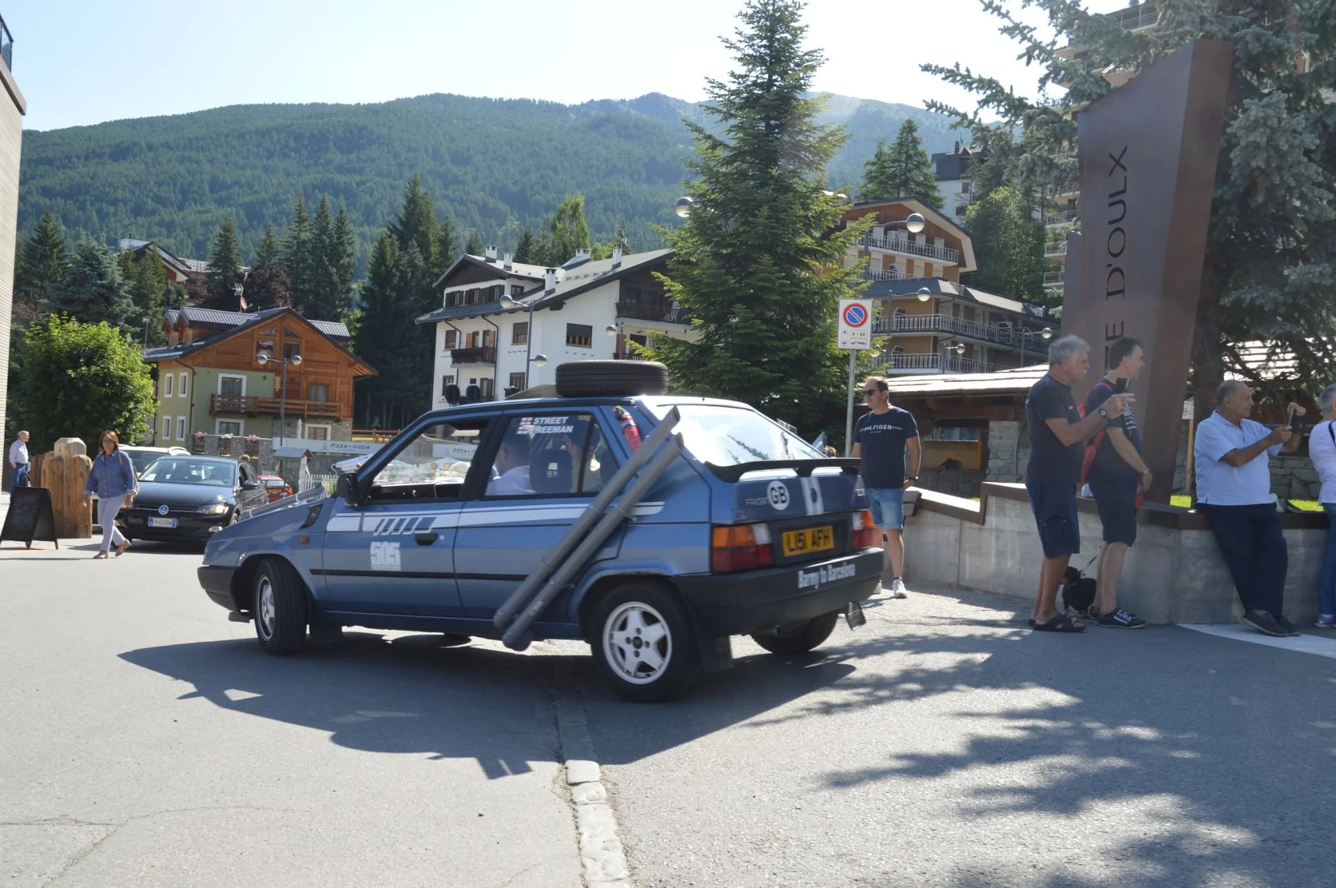 A vintage blue car with racing decals and a spare tire on the roof parked on a street in a mountain town, with people standing and walking nearby, surrounded by trees and multi-story buildings, and a large sign that reads 'Doux' in the background.
