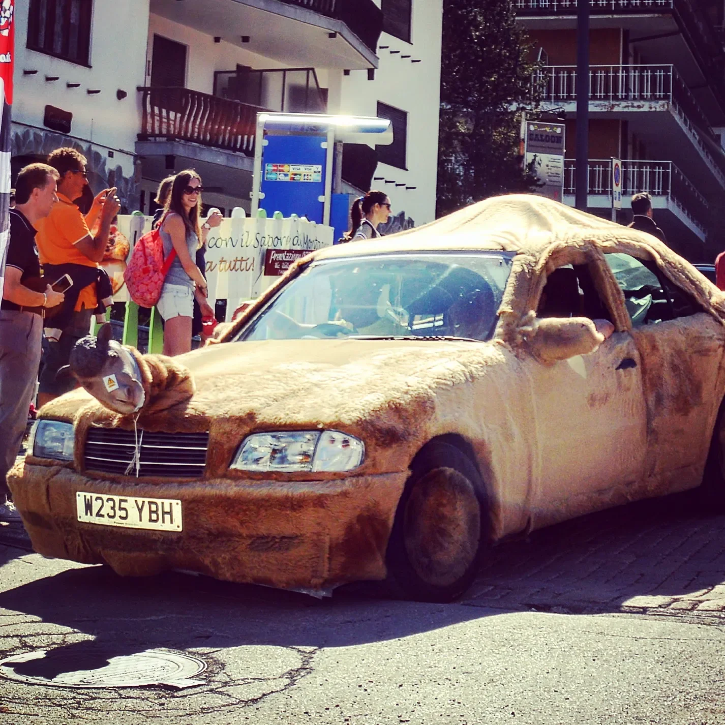 A car covered in a fake giraffe-like fur at a street event, with people and buildings in the background.