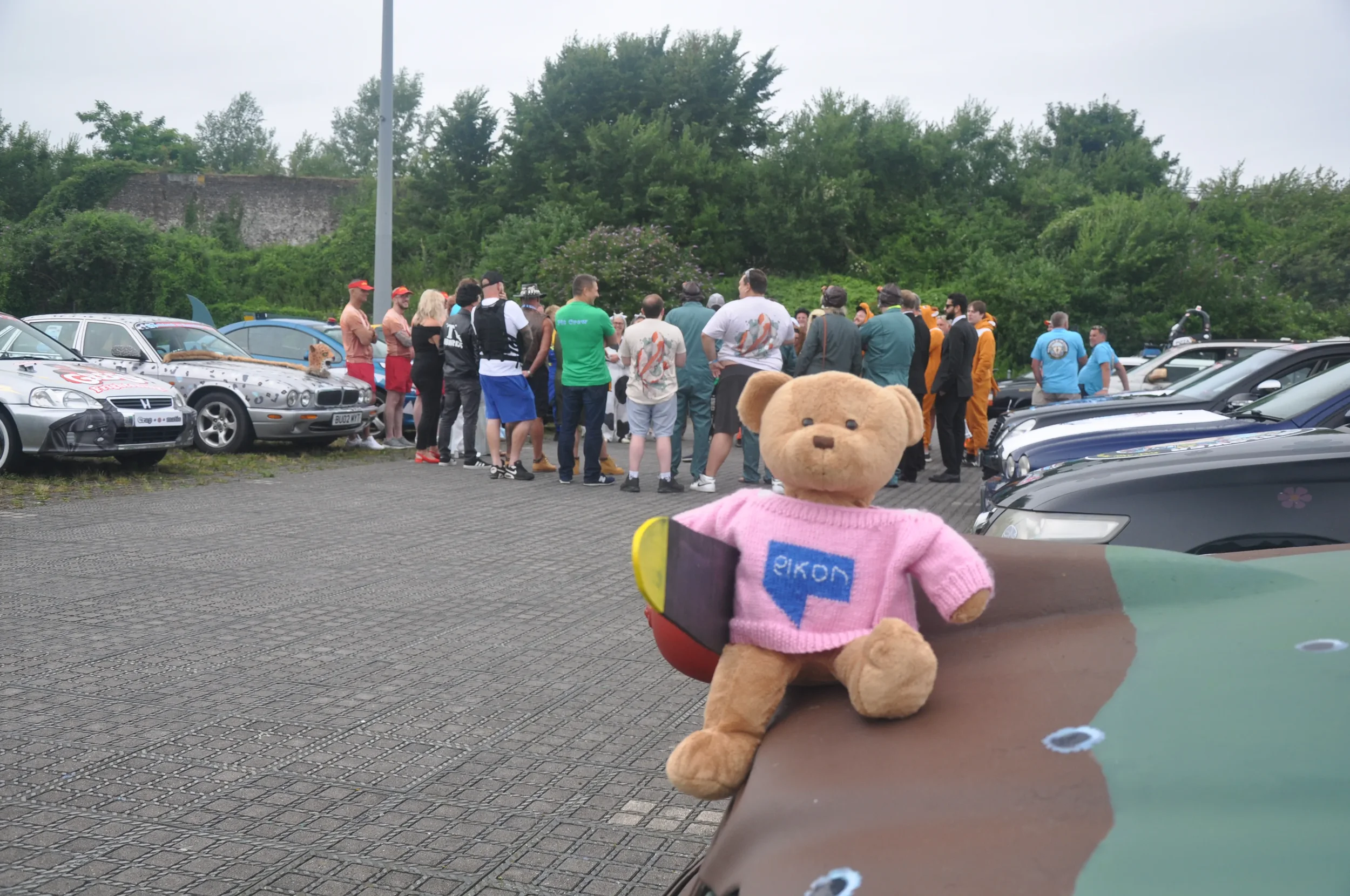 A group of people gathered outdoors in a parking lot with cars and greenery in the background. In the foreground, there is a teddy bear wearing a pink sweater with the logo 'Nokia,' placed on the surface of a painted vehicle.
