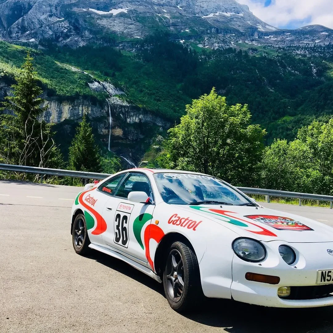 A white racing car with the number 36 and Castrol branding parked on a mountain road, with lush green trees and waterfalls in the background.