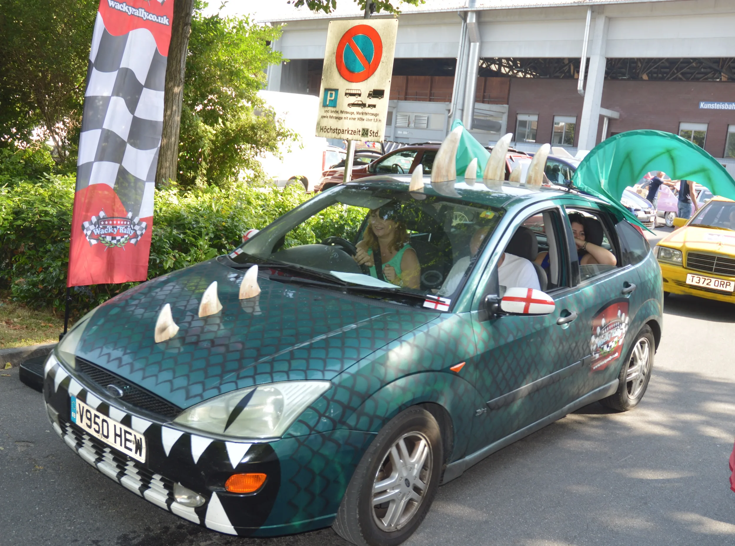 A car decorated for a race, with a scaled-green pattern, claw marks on the front, and fake horns on the roof, parked outdoors at a racing event with flags and other decorated vehicles.
