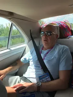 A person with short white hair, wearing sunglasses and a white shirt, sitting in the back seat of a car with a seatbelt on, with a colorful bag or blanket behind them. The car window shows greenery outside.