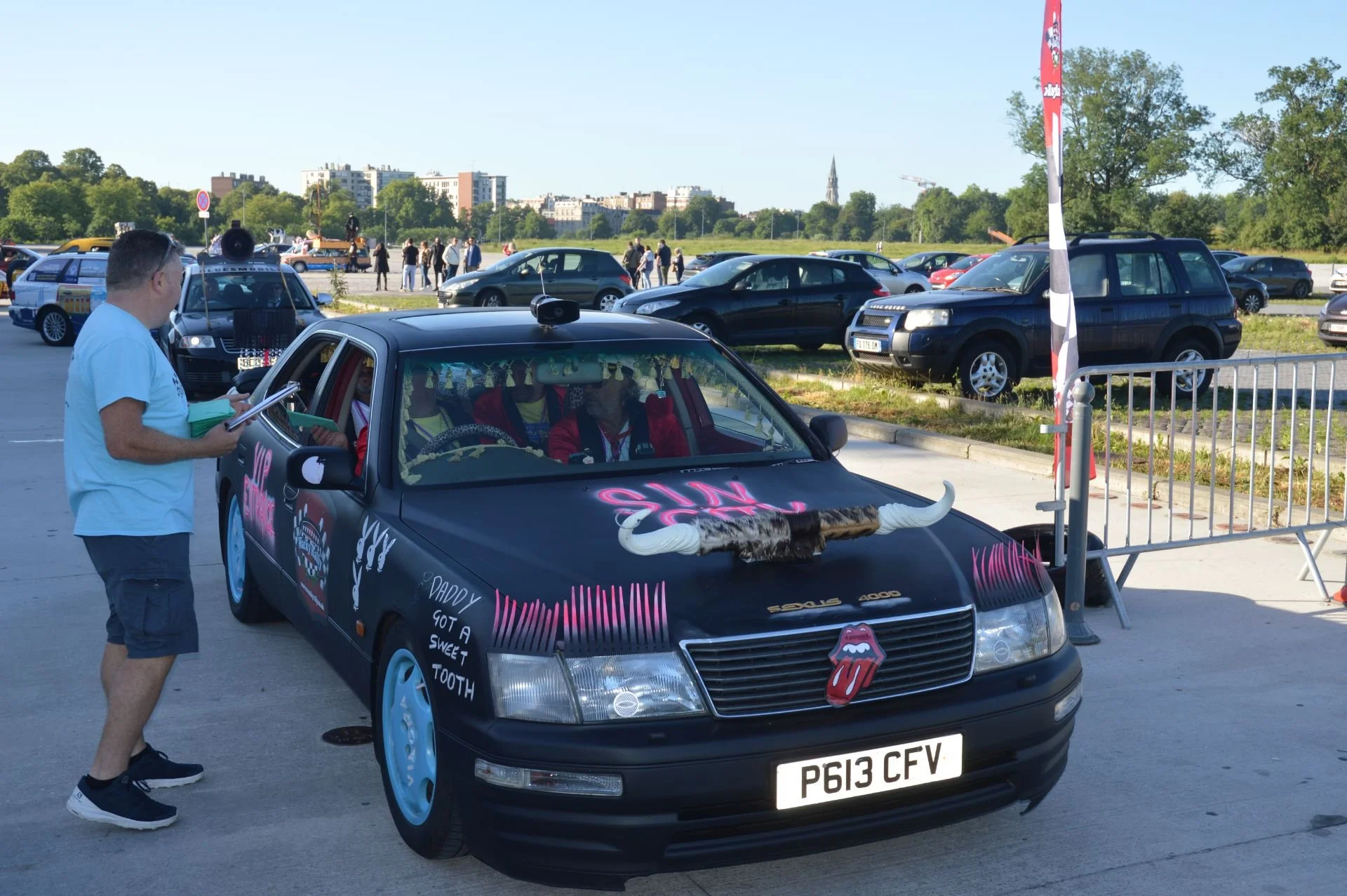 A black car decorated with colorful graffiti art, including a large fake buffalo head on the hood, a Rolling Stones tongue logo, and words like 'Sun Shine'. People are gathered around the car in an outdoor parking lot with many other vehicles and tre