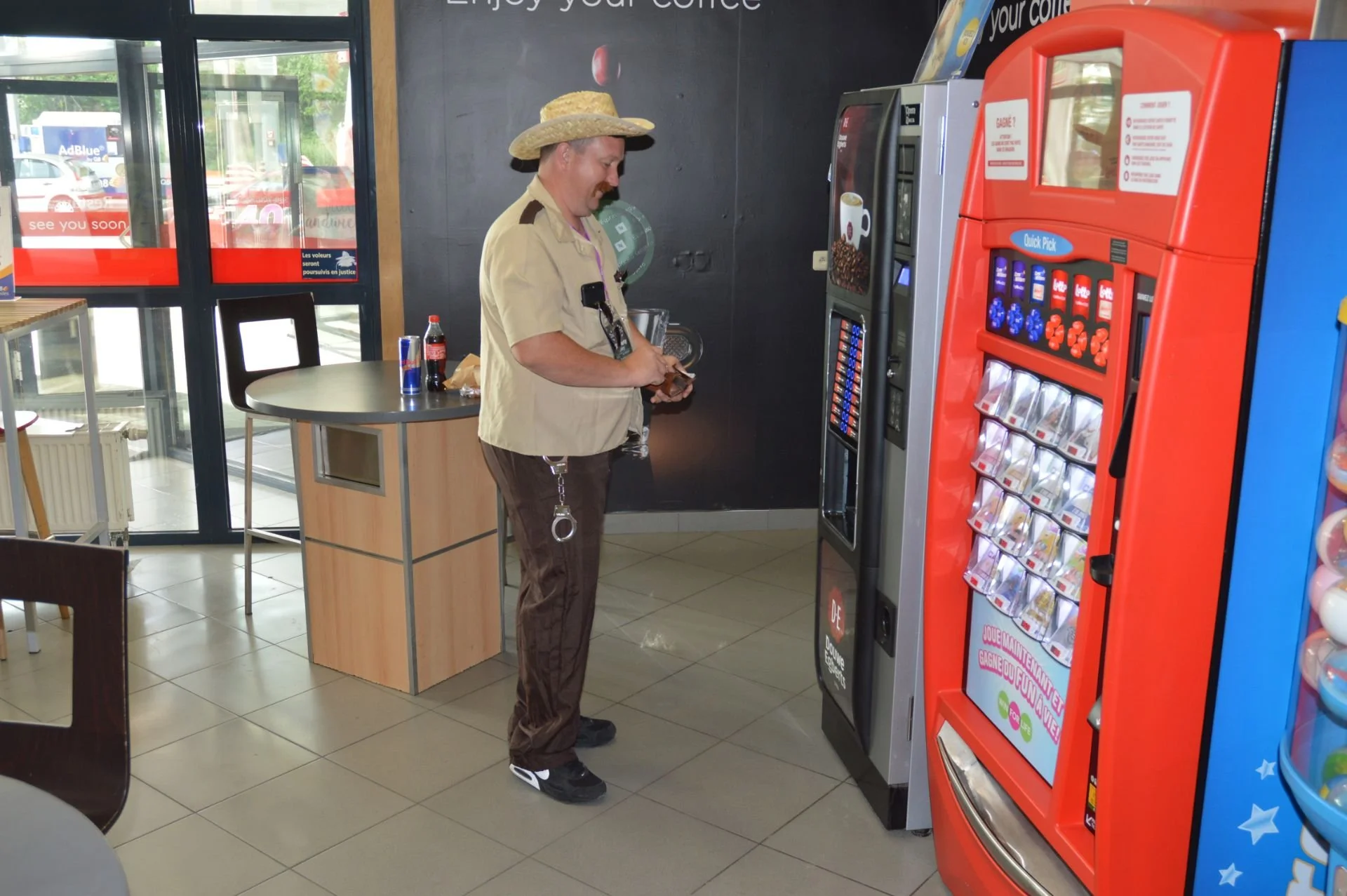 A man dressed as a security guard in a café, standing by snack and beverage vending machines, holding a phone, near a counter with a Coca-Cola bottle and snacks, with large glass doors behind him.