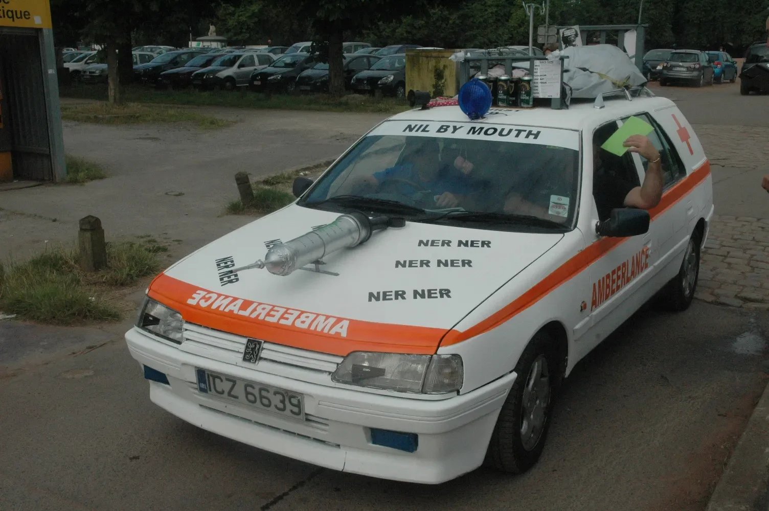 A white car with orange emergency markings and a sign that reads 'AMBER’BEER’LANCE.' The car has black text on the hood with repeated 'NER NER' and 'NER NER' and other words with misspellings like 'NIL BY MOUTH.' The car's roof is fitted with lots of