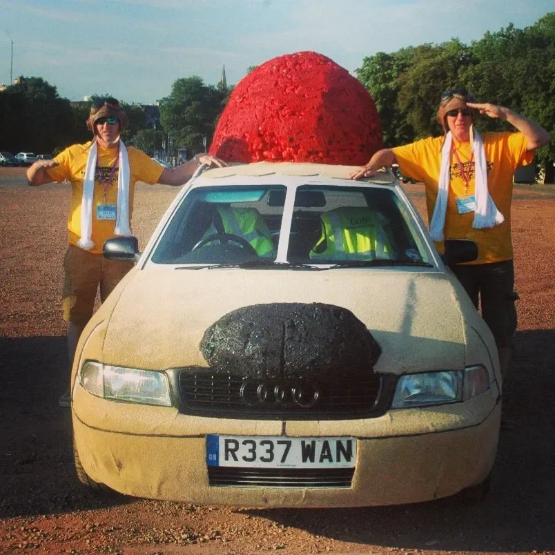Two women in yellow shirts and sunglasses stand next to a beige Audi car with a large red ball on the roof and a black sheep sculpture on the hood, parked on a gravel lot with trees in the background.