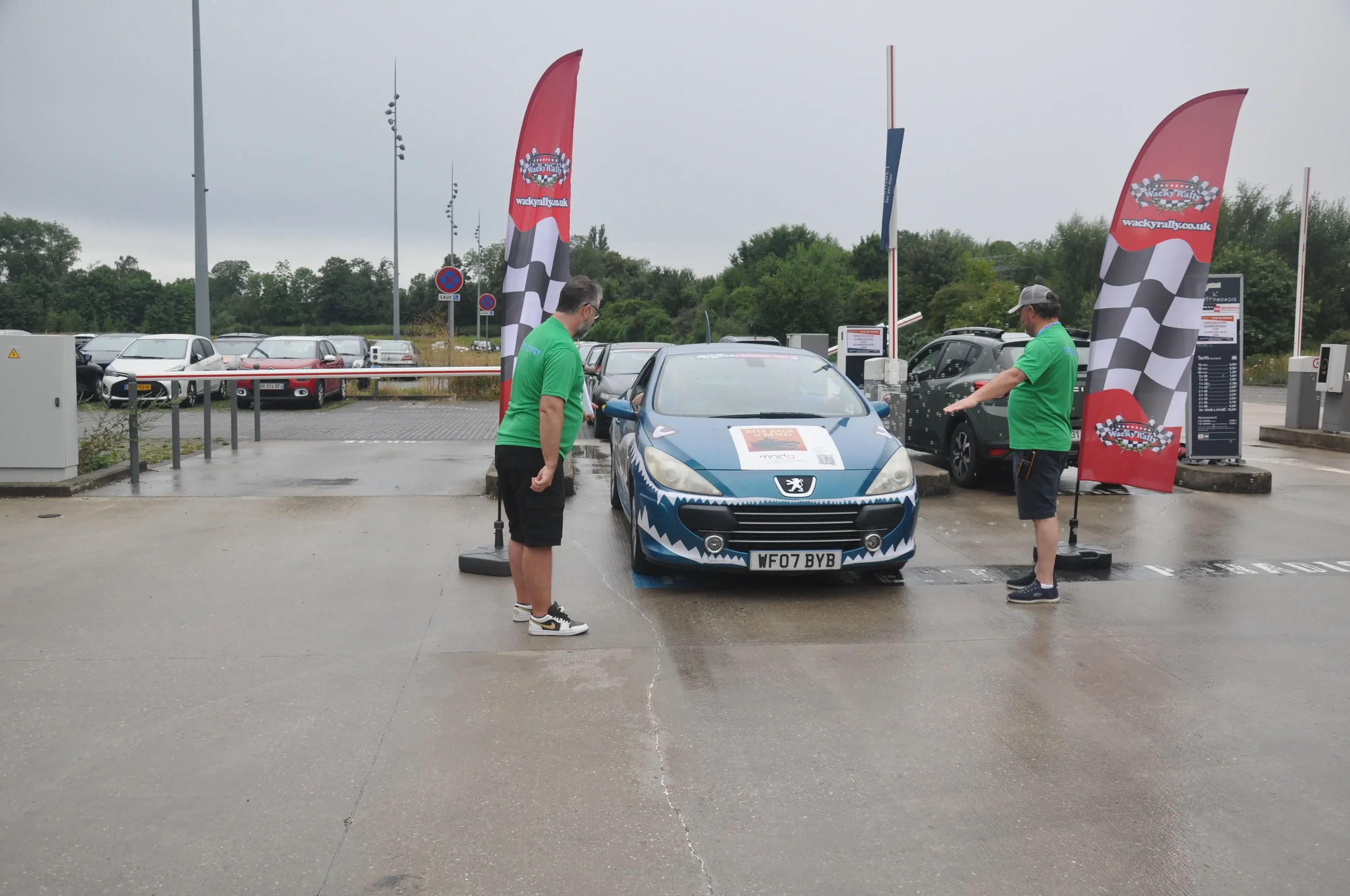 Two men in green shirts unloading a blue sports car with dragon face graphic onto a race track during a rally event at a parking lot on a rainy day, with banners and tents in the background.