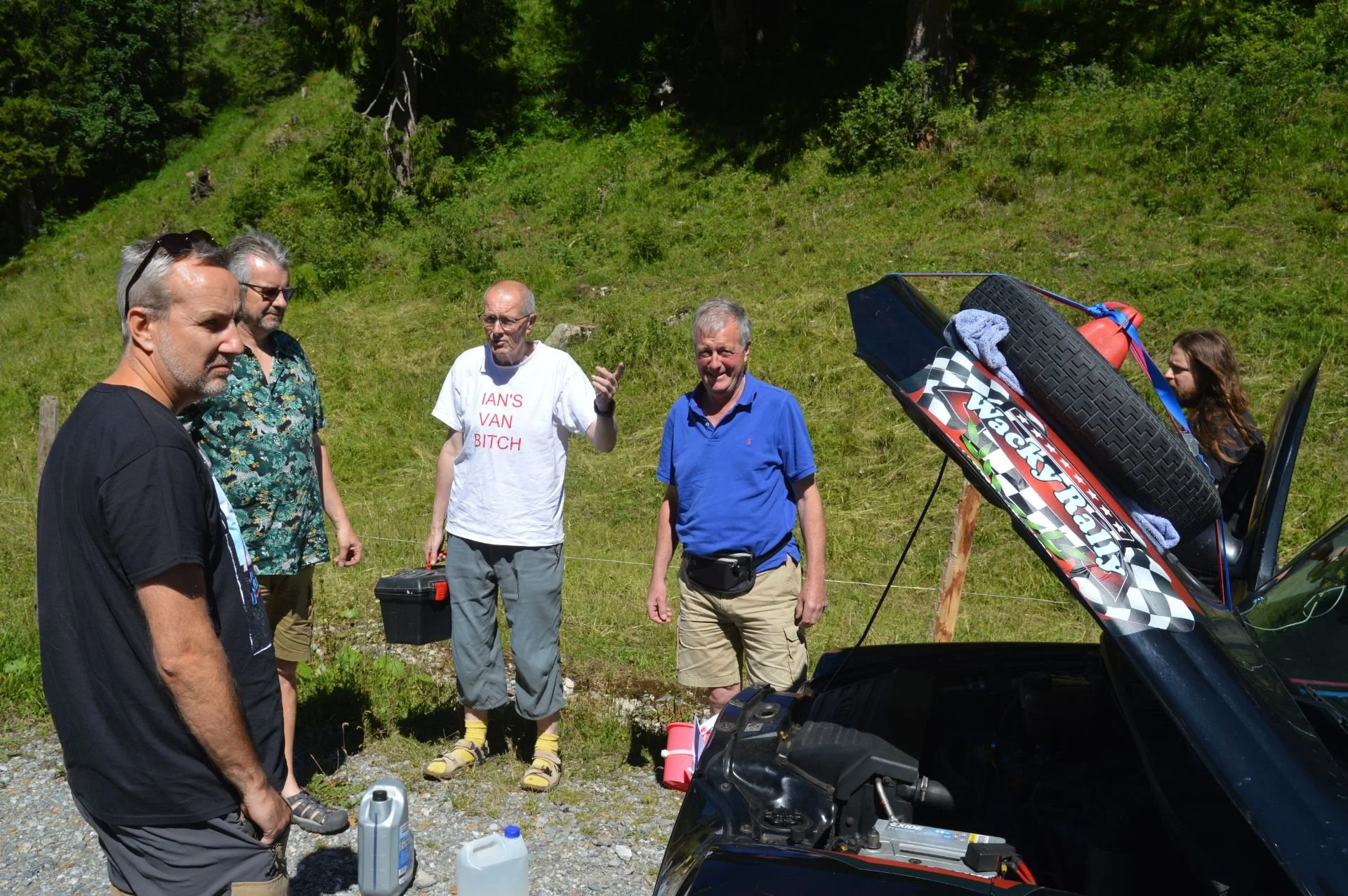 Group of five men standing outdoors near a black vehicle with its hood open, in a grassy, wooded area with hills in the background. One man in a white T-shirt with red text is gesturing. The scene appears to be a casual outdoor gathering on a sunny d