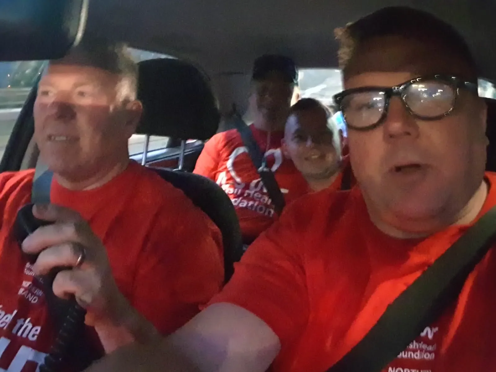 Four men wearing red shirts sitting inside a car, with the driver holding the steering wheel and a woman smiling in the back seat.