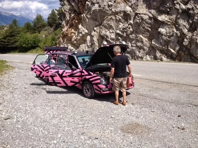 A man with gray hair, wearing a black shirt and beige shorts, is standing next to a pink car with black geometric patterns. The car's hood and trunk are open, and the man is looking into the engine. The car is parked beside a rocky hillside and a pav