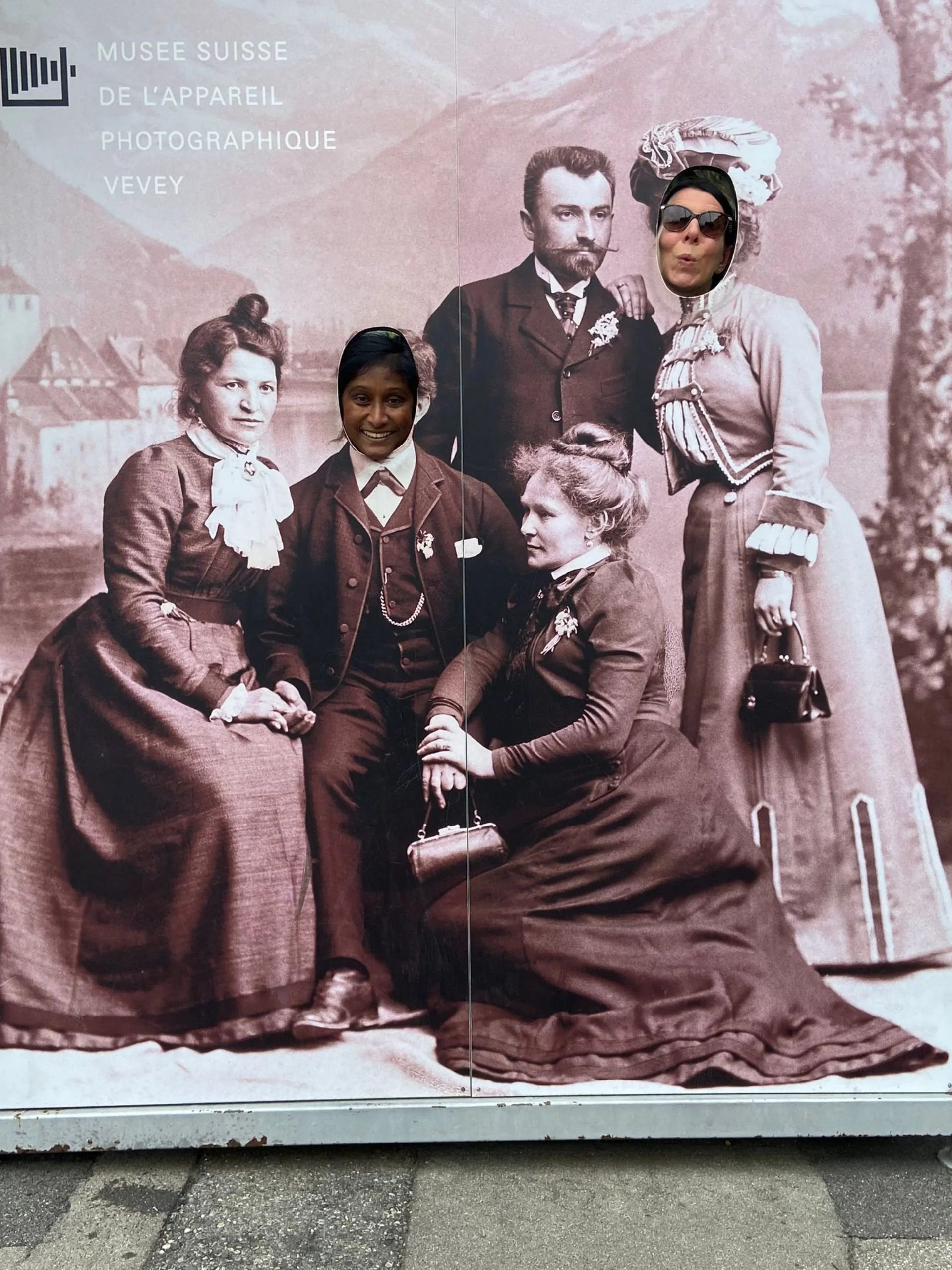 An outdoor sign at the Swiss Museum of Photography Vevey features a vintage black-and-white photo of five people, with two faces replaced by modern people wearing sunglasses and a hat.