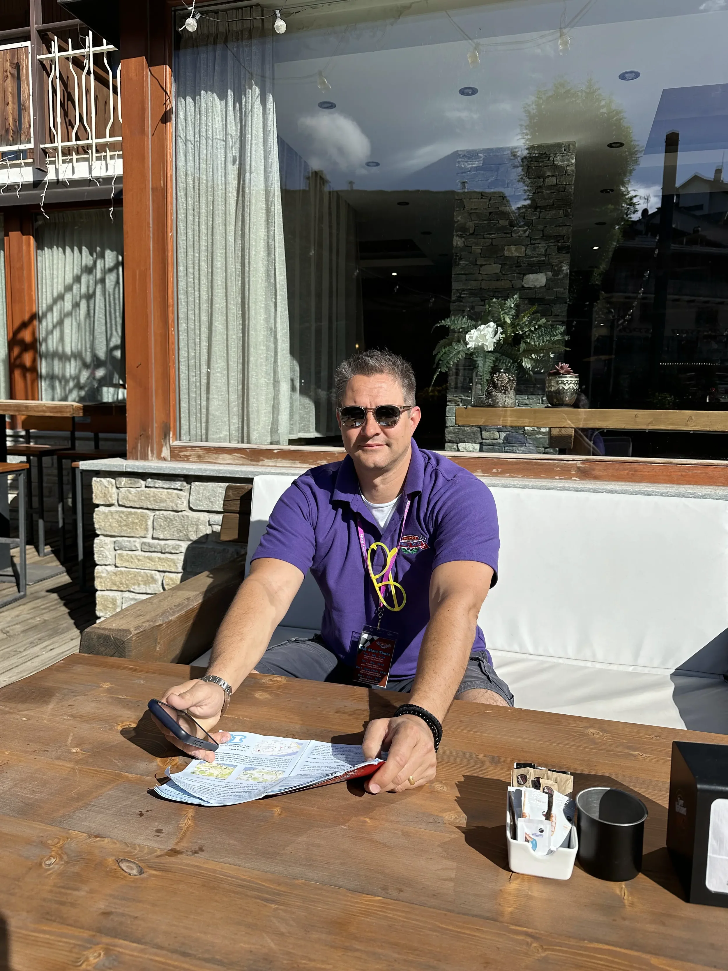 Man in sunglasses sitting at an outdoor wooden table, holding a smartphone, with a restaurant window behind him.