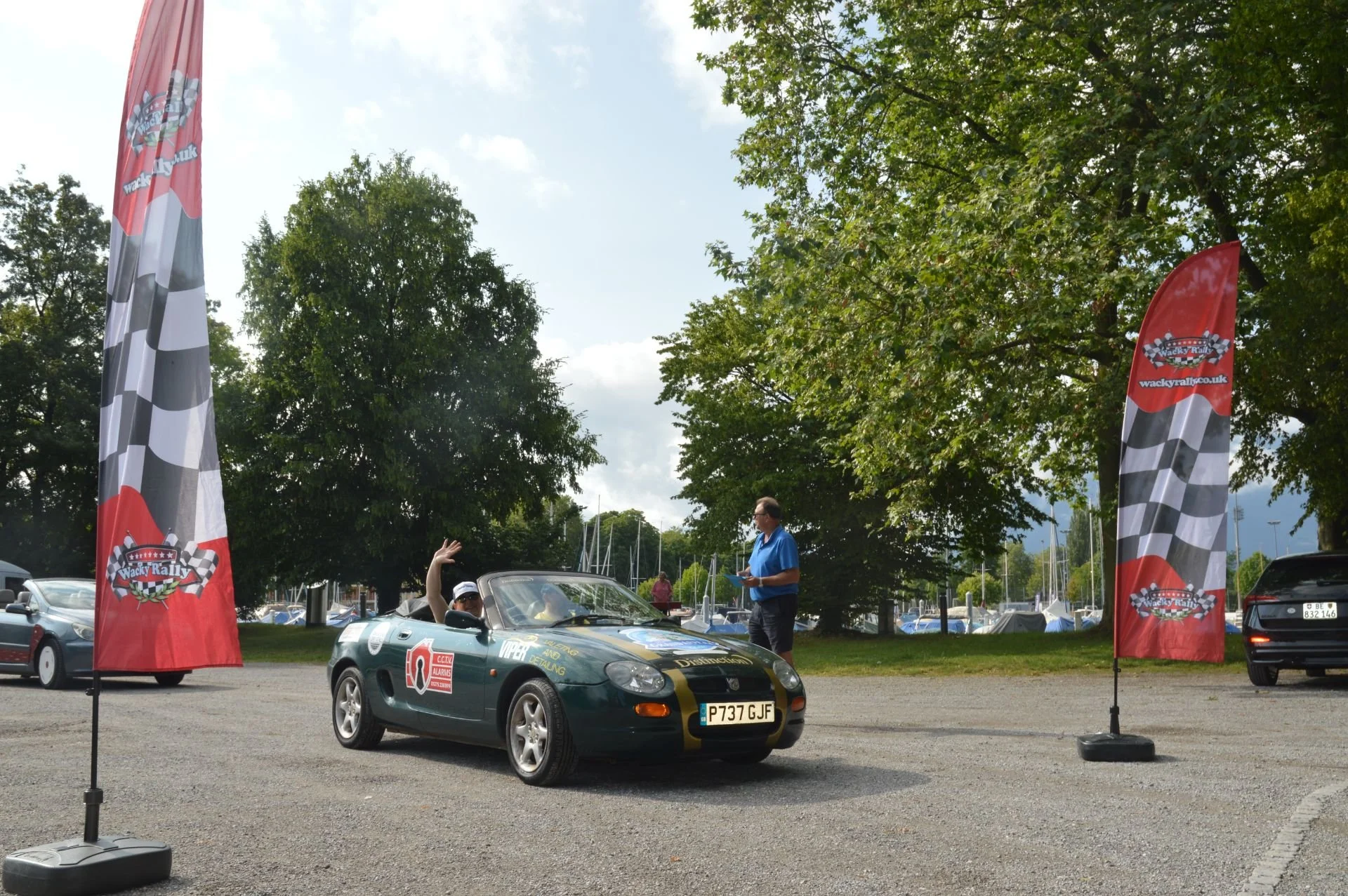 A green convertible car with racing decals and a license plate P737 GJF is parked in an outdoor lot. A person is seated inside the car waving, while another person stands nearby holding a clipboard. Two tall flags with racing checkered patterns and l