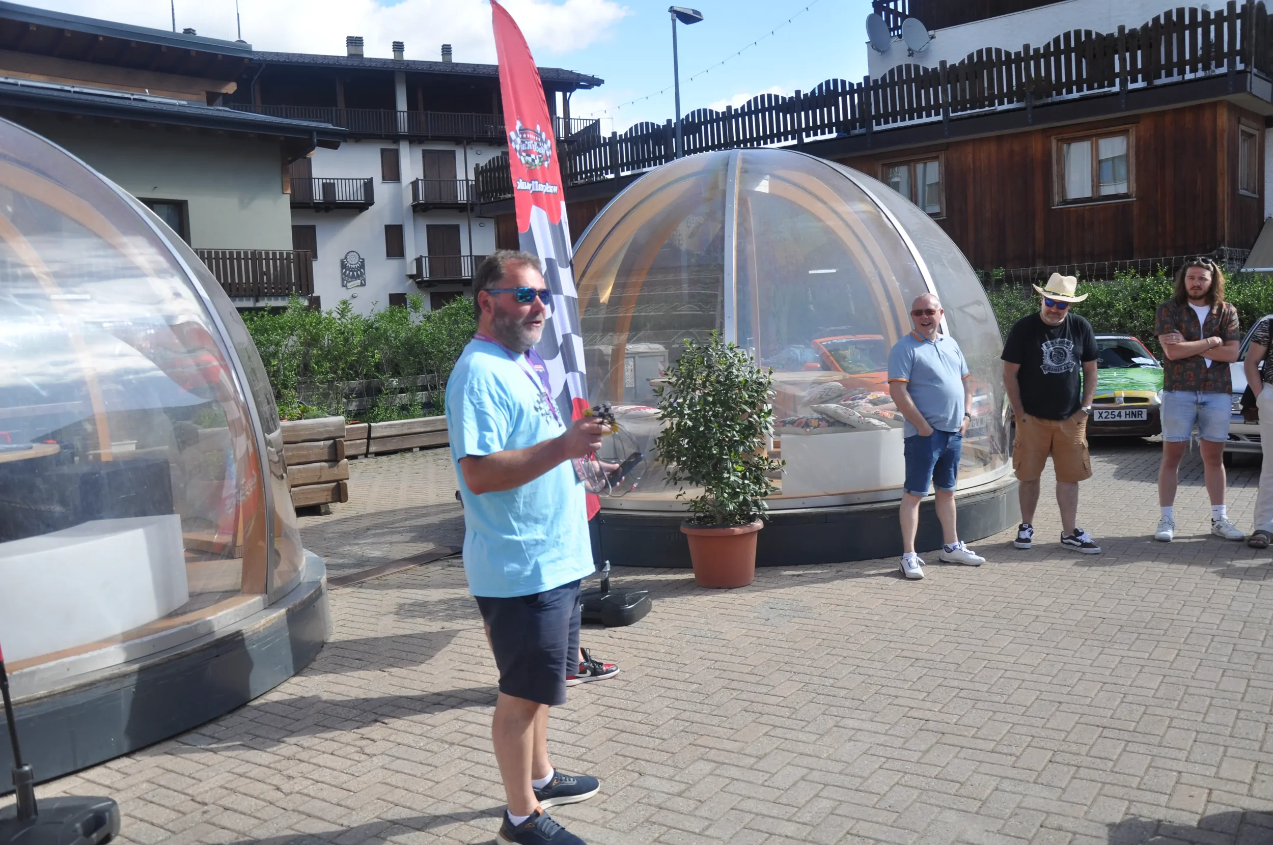 A group of men standing outdoors in front of transparent geodesic dome structures, with a man in a blue shirt holding a drone controller, and others casually talking or listening, in a paved courtyard area with residential buildings in the background