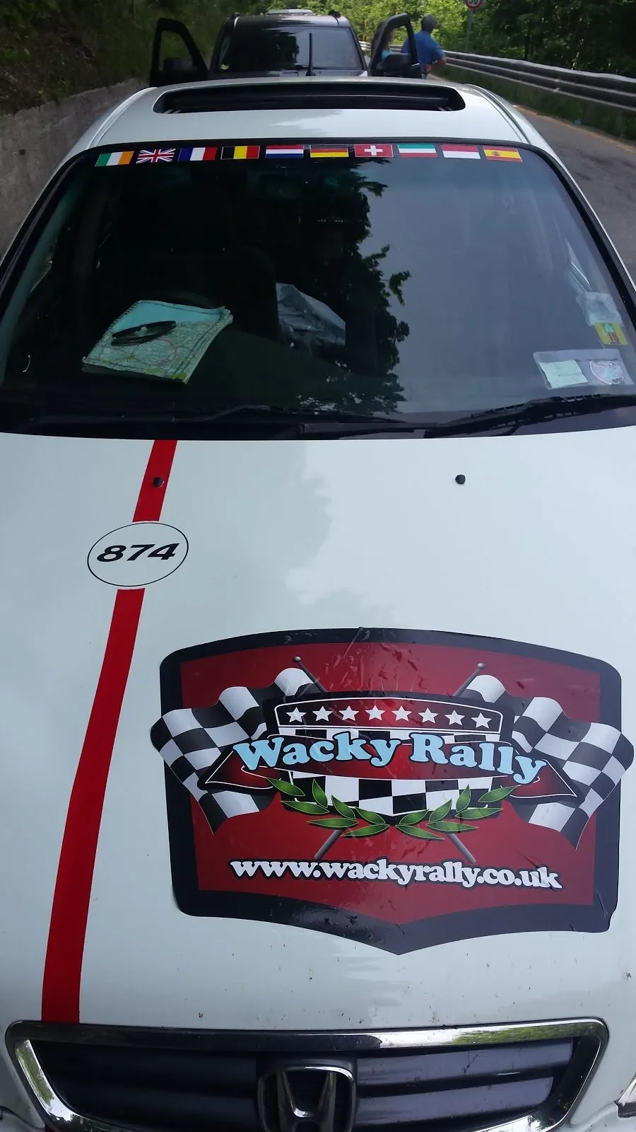 Front view of a white race car with a Wacky Rally logo, racing flags, and the website URL on the hood, parked on a road with a guardrail and trees.