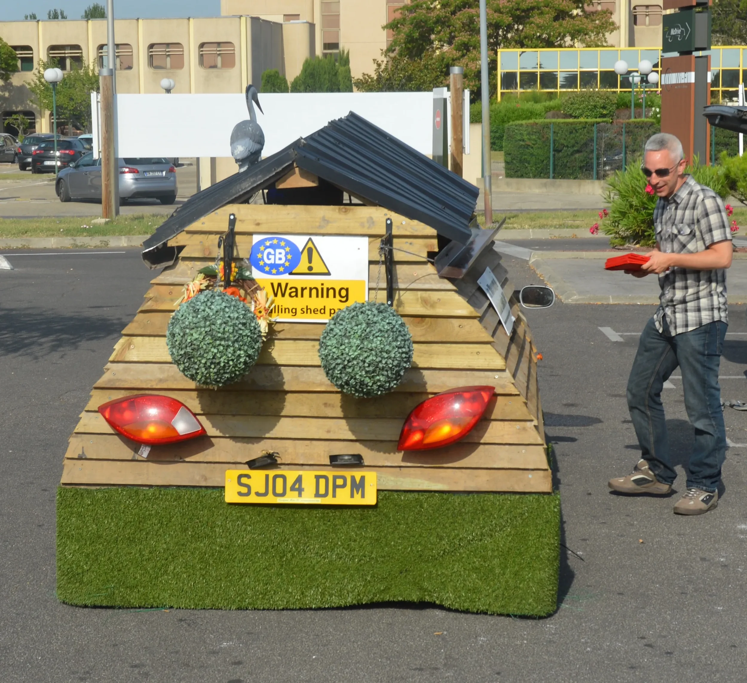 A small wooden shed or cart decorated with two round artificial bushes, a warning sign, and two red rear lights, with a license plate reading 'SJ04 DPM'. A man wearing sunglasses, a plaid shirt, and jeans is standing nearby holding a red book or fold