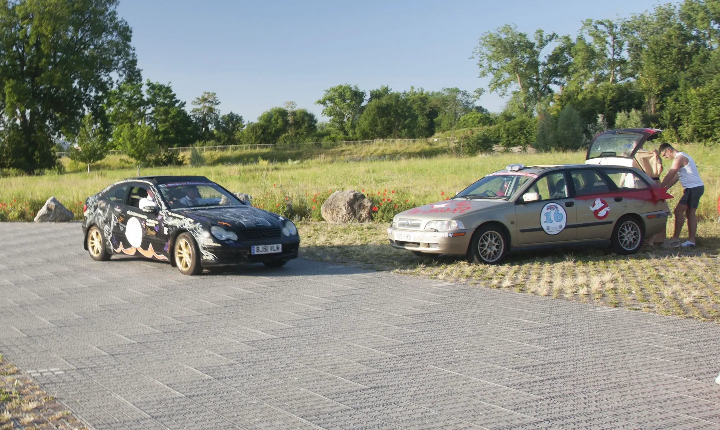 Two decorated cars parked on a gravel lot with a man standing next to one of the cars. The background features green trees and grassy area with rocks.