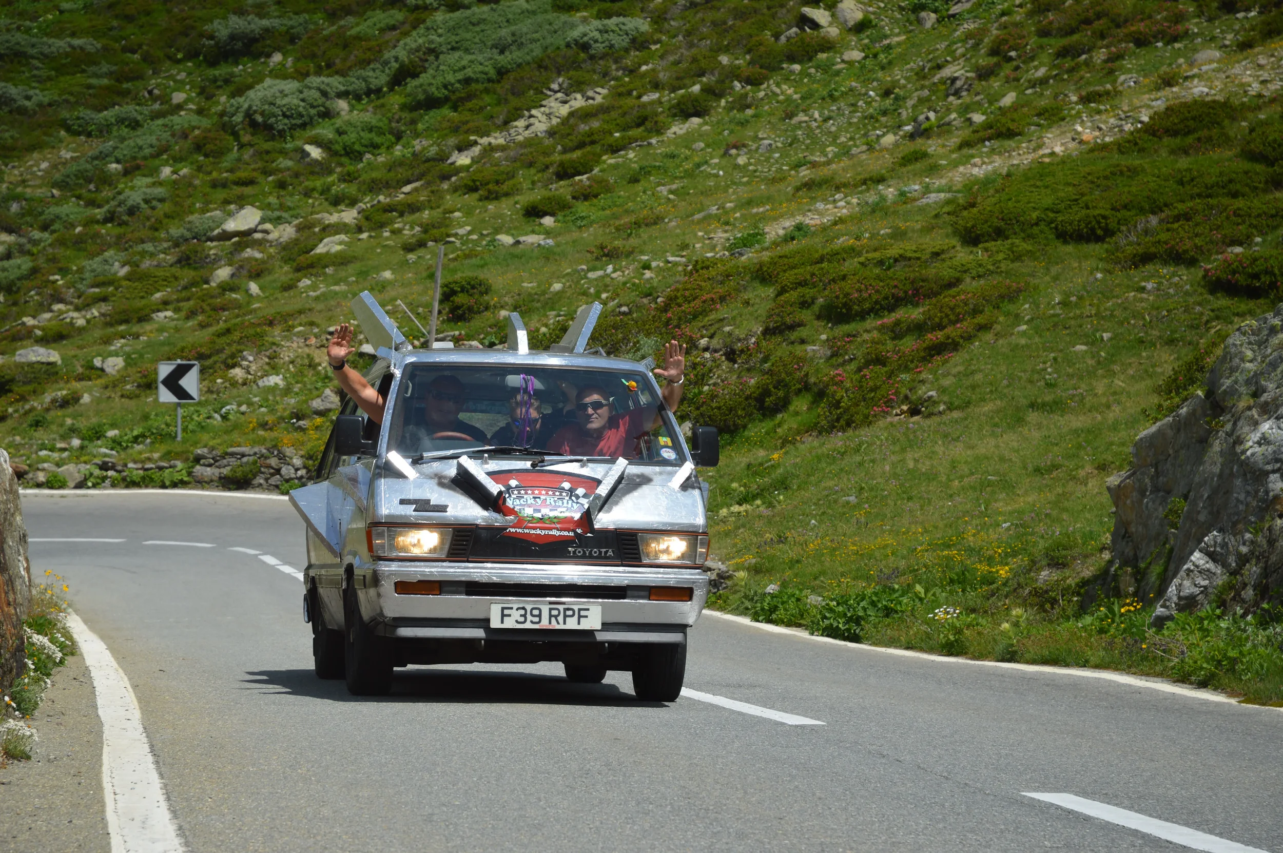 A silver car decorated with stickers and a sign on the front driving on a mountain road with two people inside, one waving with both hands and the other wearing sunglasses. The road is curvy with green grass and rocks on the sides.