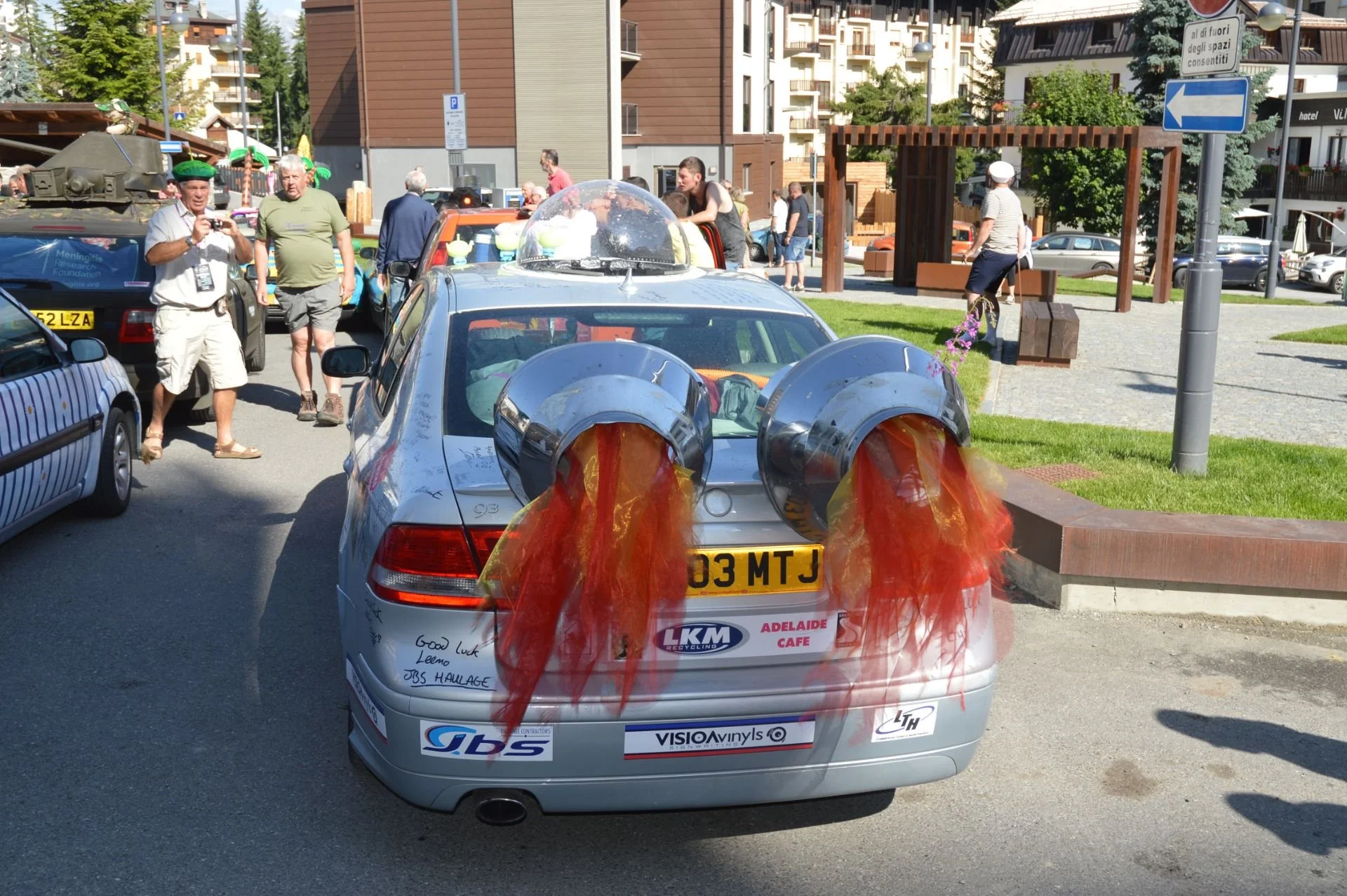 A silver car with two tuba-like loudspeakers on its trunk, emitting red and yellow fabric streamers, at an outdoor event with people walking and taking photos.