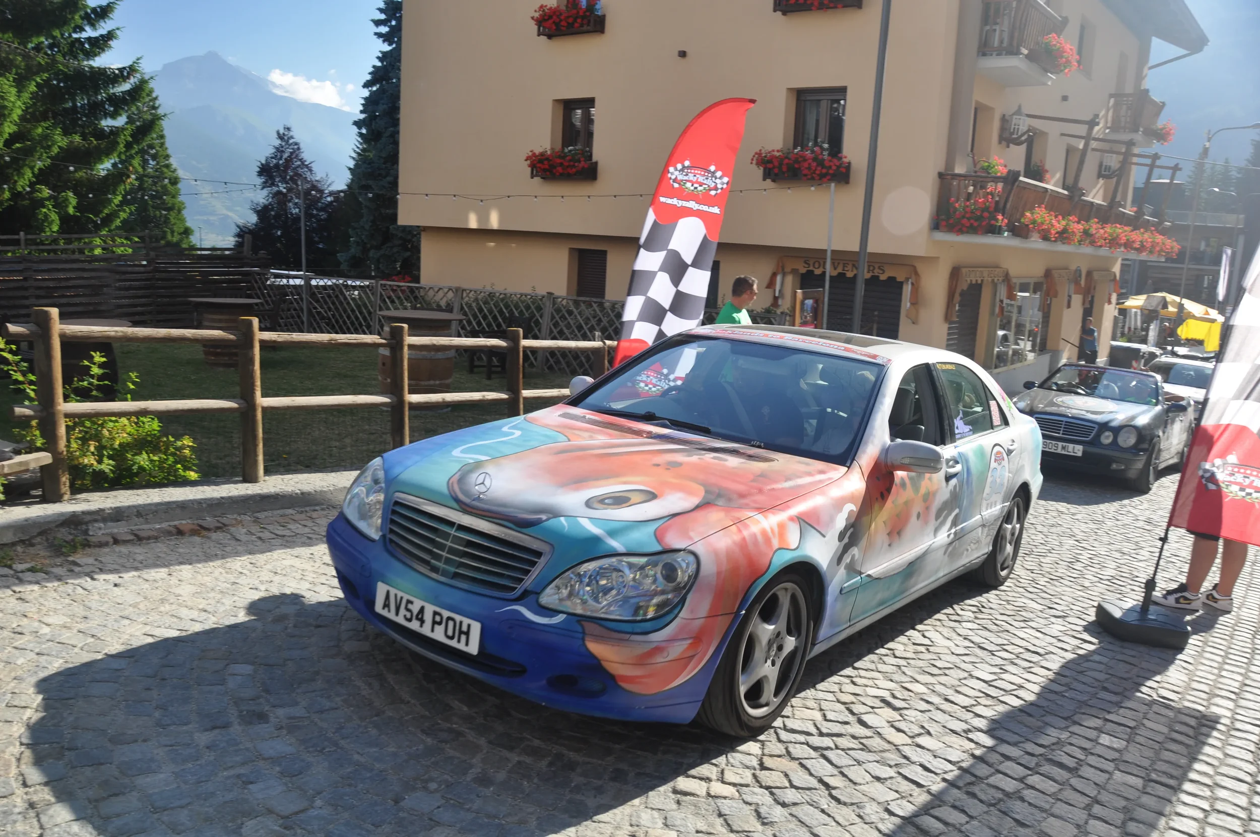 A Mercedes-Benz car with a colorful painted design parked on a cobblestone street during a car show event, with a checkered flag-shaped banner nearby and another black car behind it.