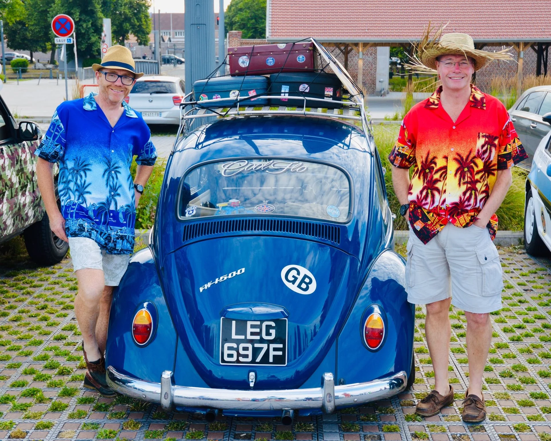 Two men in colorful summer shirts and shorts standing on either side of a vintage blue Volkswagen Beetle car with a roof rack loaded with luggage, in an outdoor parking lot.