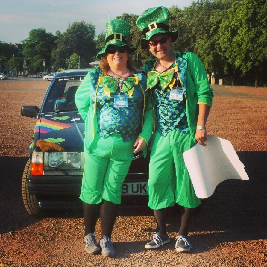 Two people dressed in St. Patrick's Day costumes standing in front of a black car with a rainbow design on the hood and shamrocks, with a parking lot and trees in the background.
