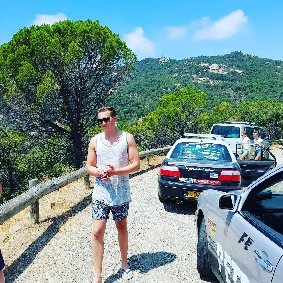 A young woman in sunglasses and casual summer clothes standing on a gravel mountain road with green trees and hills in the background. There are three cars parked nearby, including a police car and two other vehicles, with two people near the white v