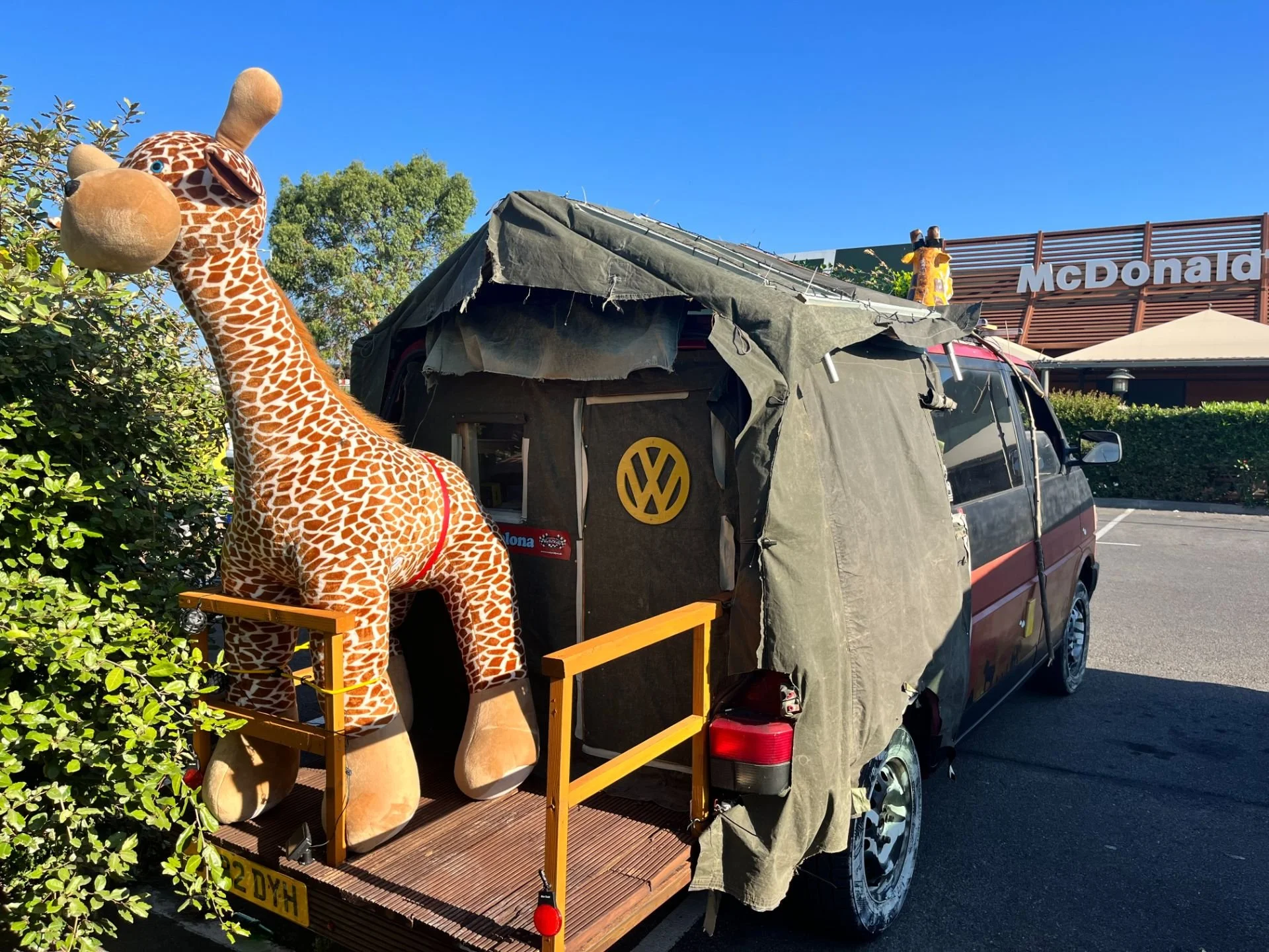 A plush giraffe toy mounted on a miniature vehicle with a VW logo, parked outside a McDonald's restaurant.