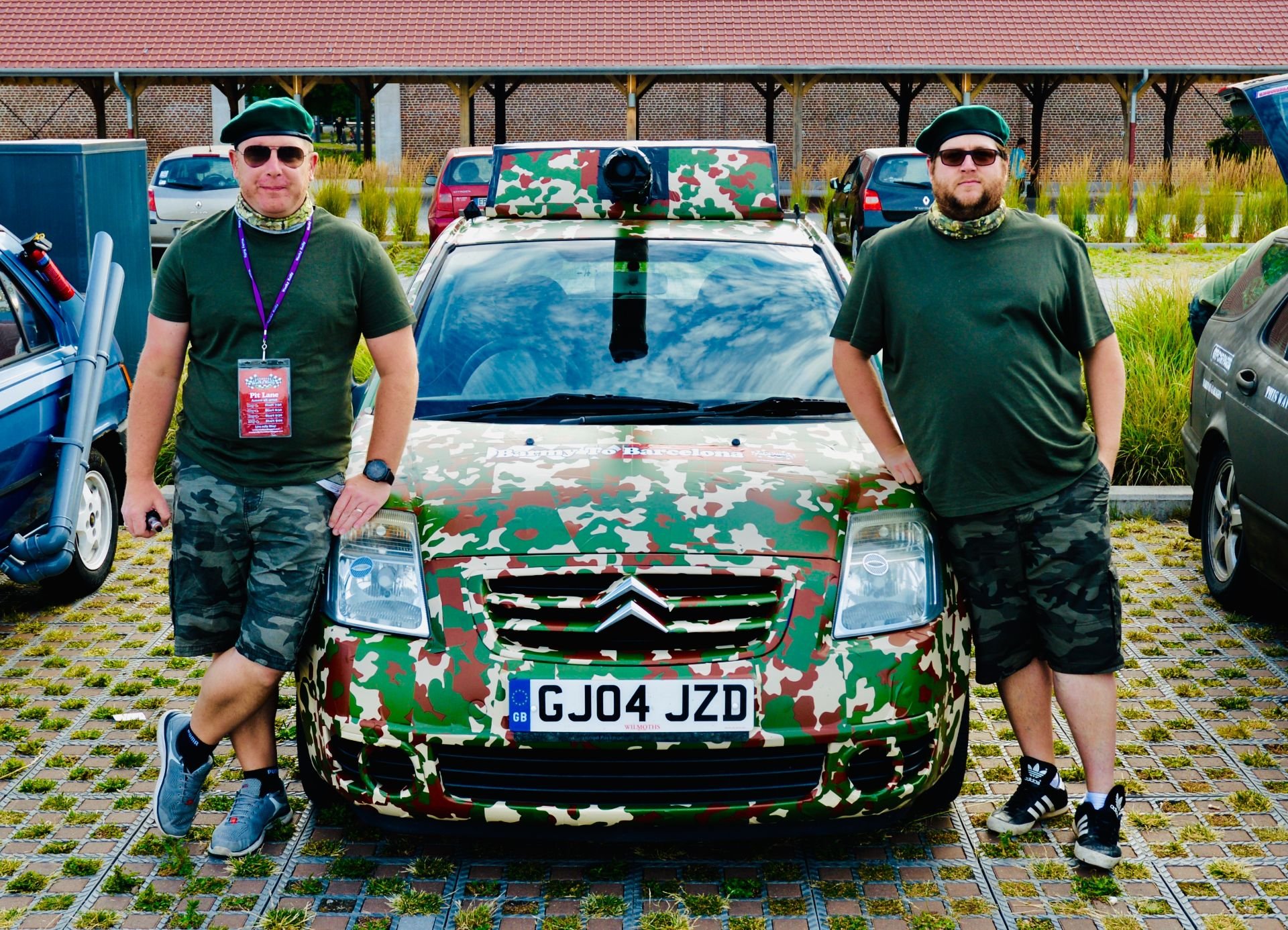 Two men wearing green shirts, camouflage shorts, and green berets standing beside a camouflaged Citroën car with a camouflage roof box, in a parking lot with other cars and a brick building in the background.
