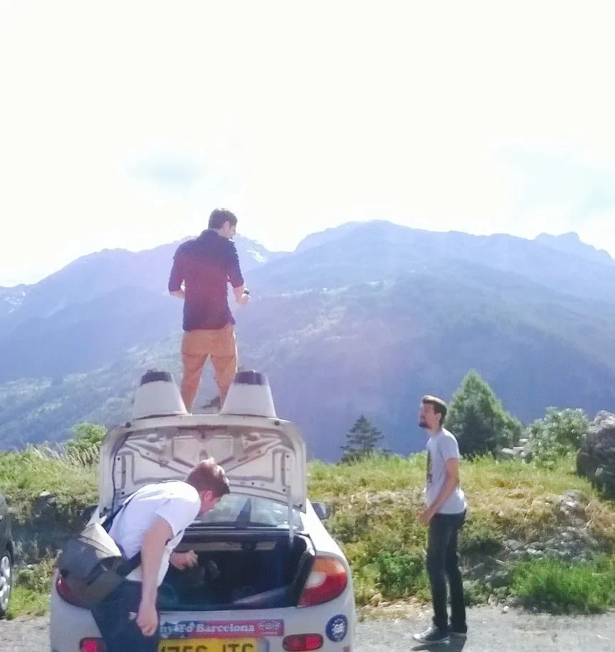 Three men and a woman in outdoor clothing near a white car with the trunk open. One man is standing on the car's roof, another is leaning into the trunk, and the third is standing on the ground nearby. The scene is set against a backdrop of mountains