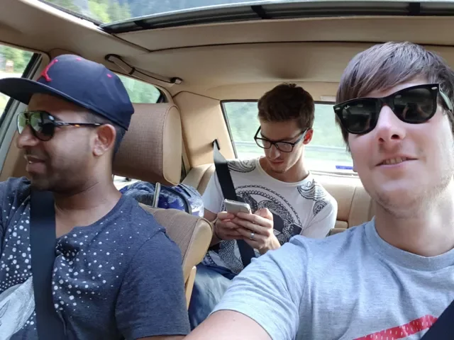 Three young men are sitting in the back seat of a car, with the man in the front taking a selfie. The man on the left is wearing a navy baseball cap, sunglasses, and a navy shirt with white polka dots. The man in the middle has brown hair, glasses, a