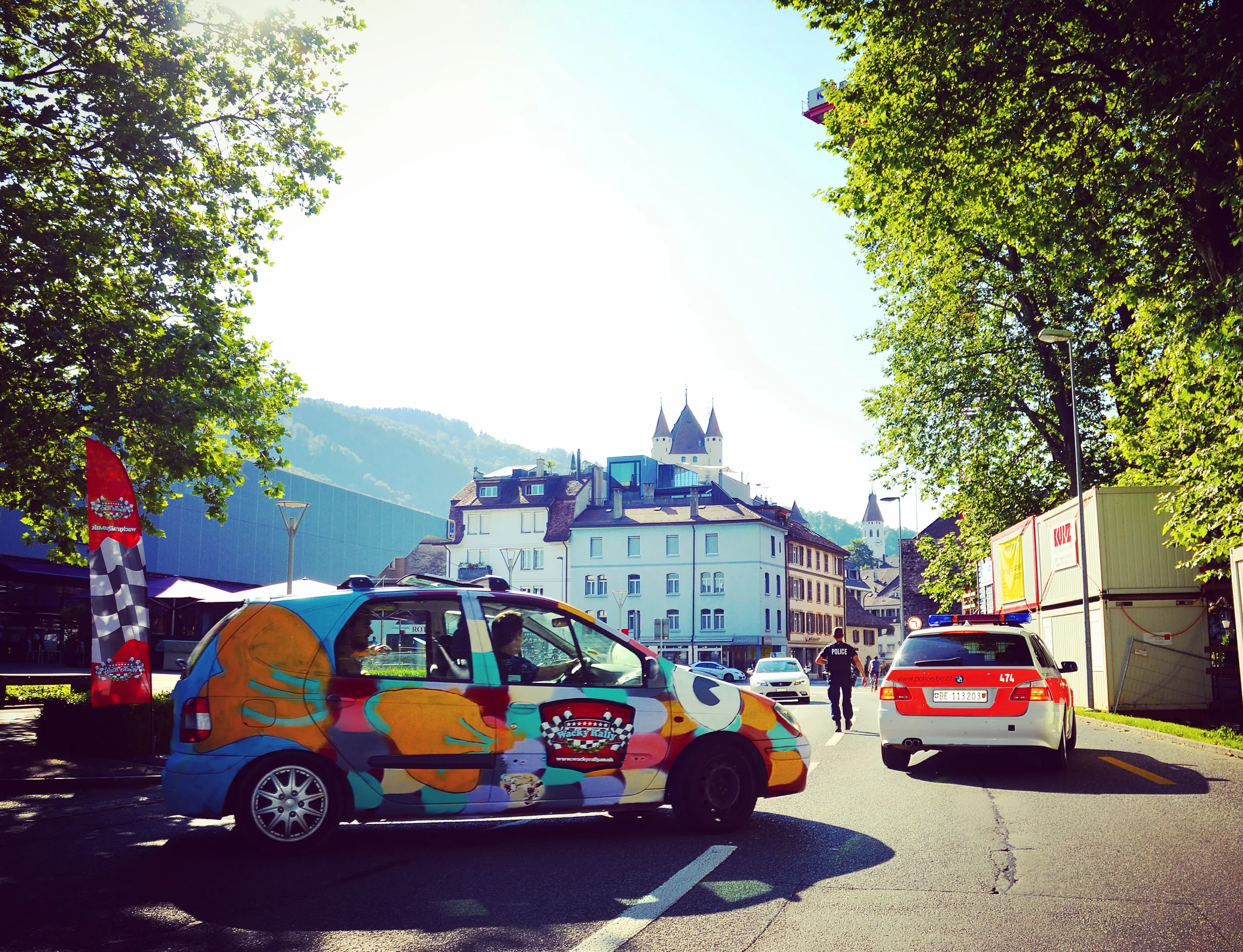 A colorful van with a panda, orange art, and checkered pattern on it is parked, with a police car beside it on a sunny street lined with trees and buildings, including a church with towers, in the background.