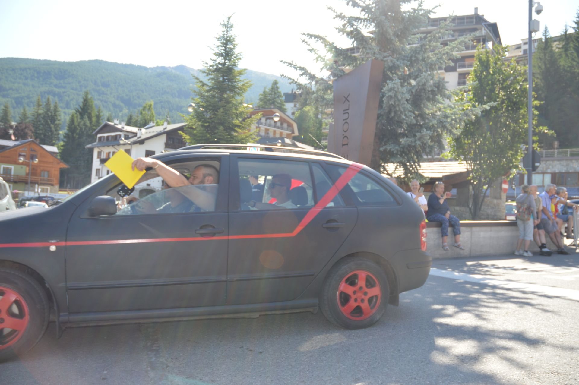 Black car with red rims parked on the street, with a man inside raising a yellow envelope or folder out of the window. In the background, there are people sitting on a stone ledge, trees, buildings, and a sign that reads 'D'OLUX', with mountains in t