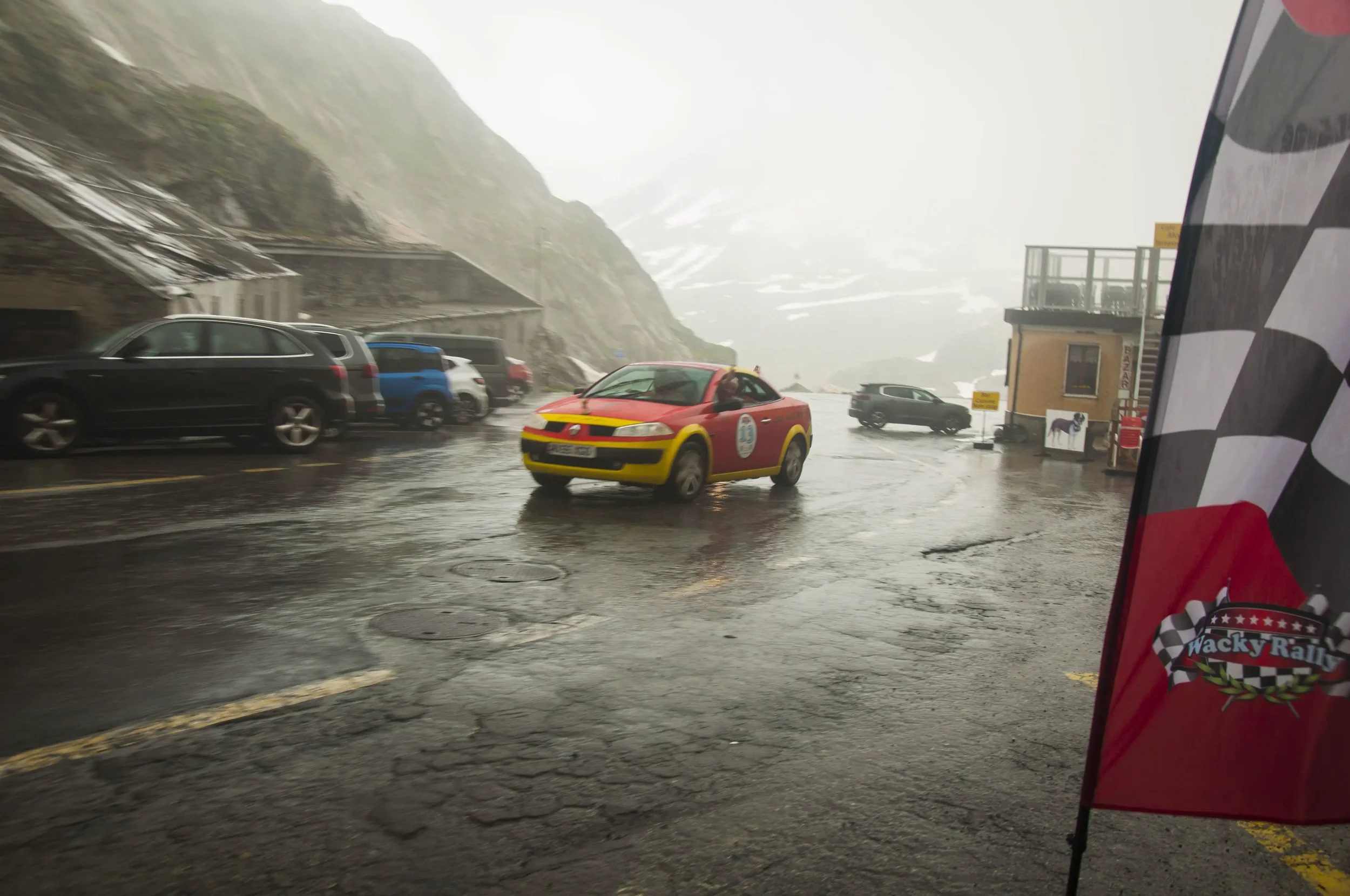 Rainy mountain roadside with parked cars, a red and yellow race car, and a rally flag with a checkered pattern and the words "Wacky Rally" on it.