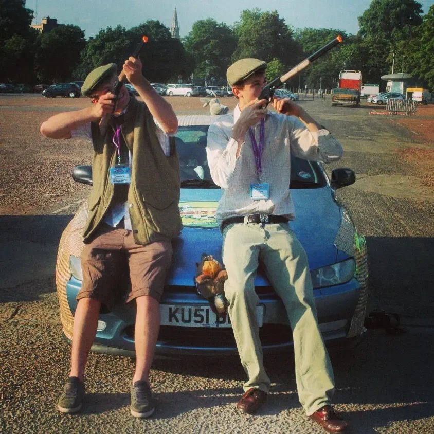 Two young men in vintage style clothing, wearing hats and sunglasses, standing on the hood of a car and aiming toy rifles into the distance in a parking lot with trees and other vehicles in the background.