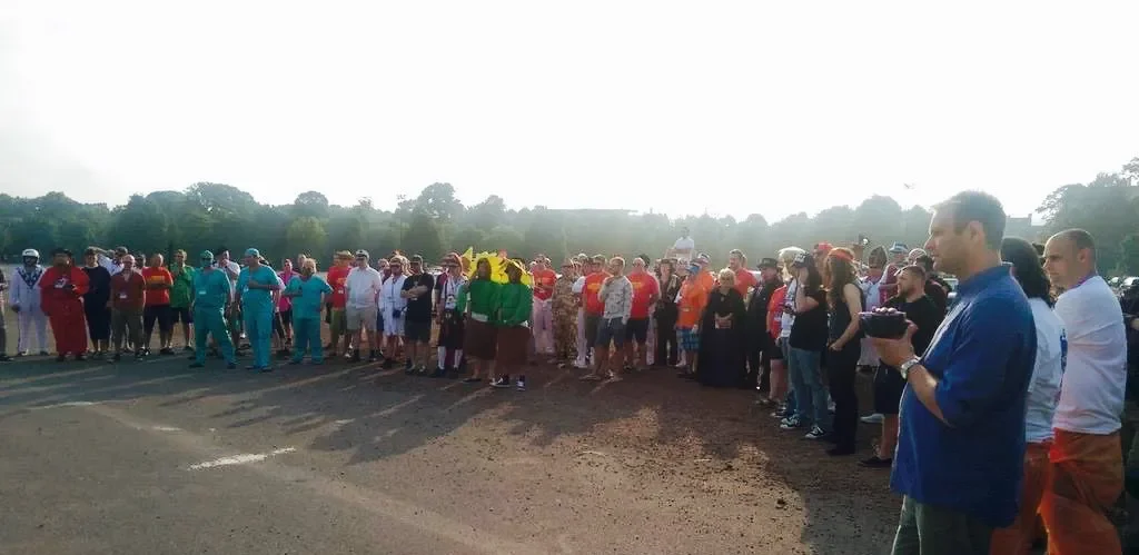 Large group of people gathered outdoors during daytime, some dressed in colorful clothing, standing on a dirt surface with trees and hills in the background.