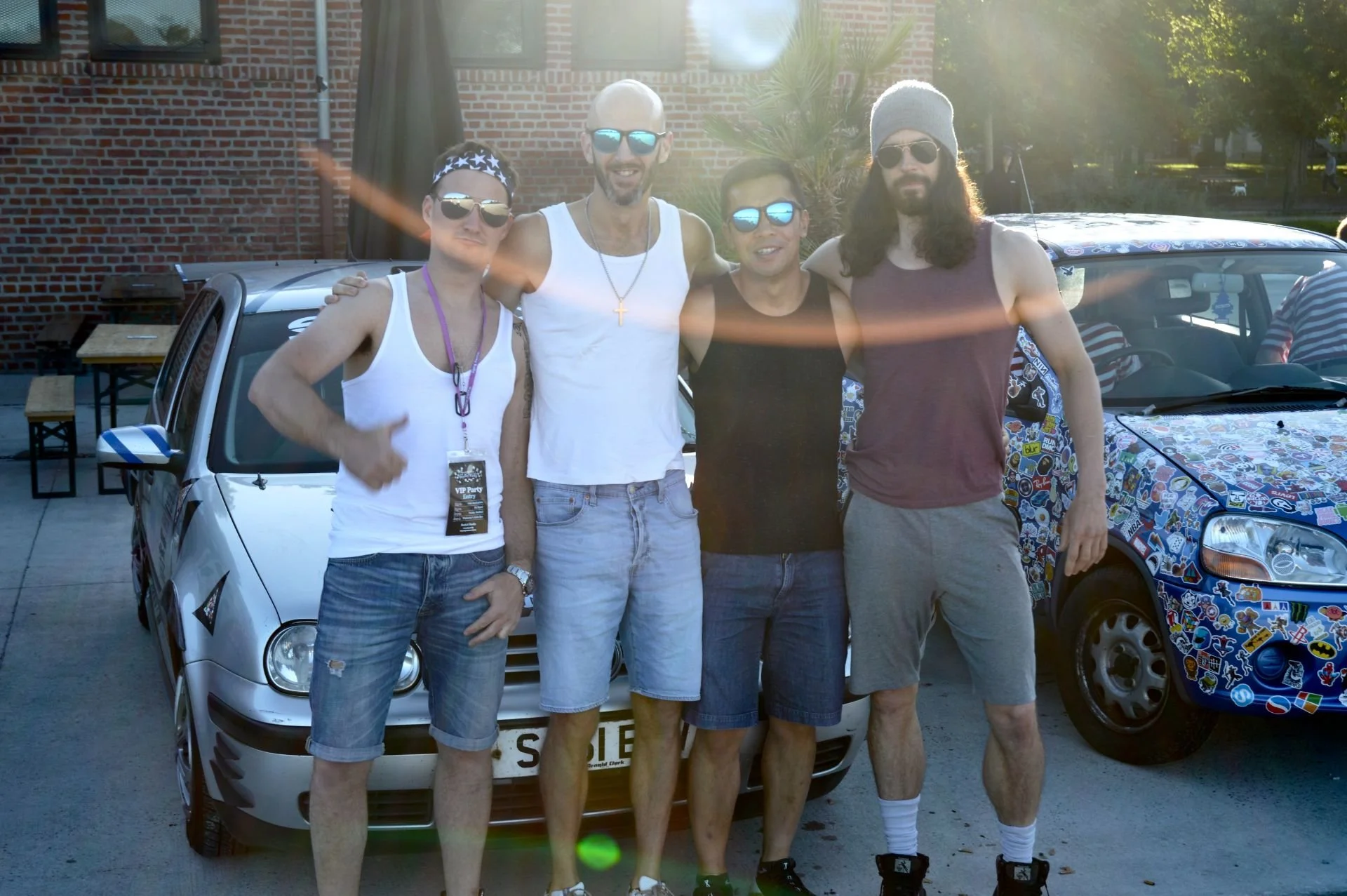 Four men standing in front of a silver car and a colorful sticker-covered car, outdoors with sunlight, brick building, trees, and tables in the background.