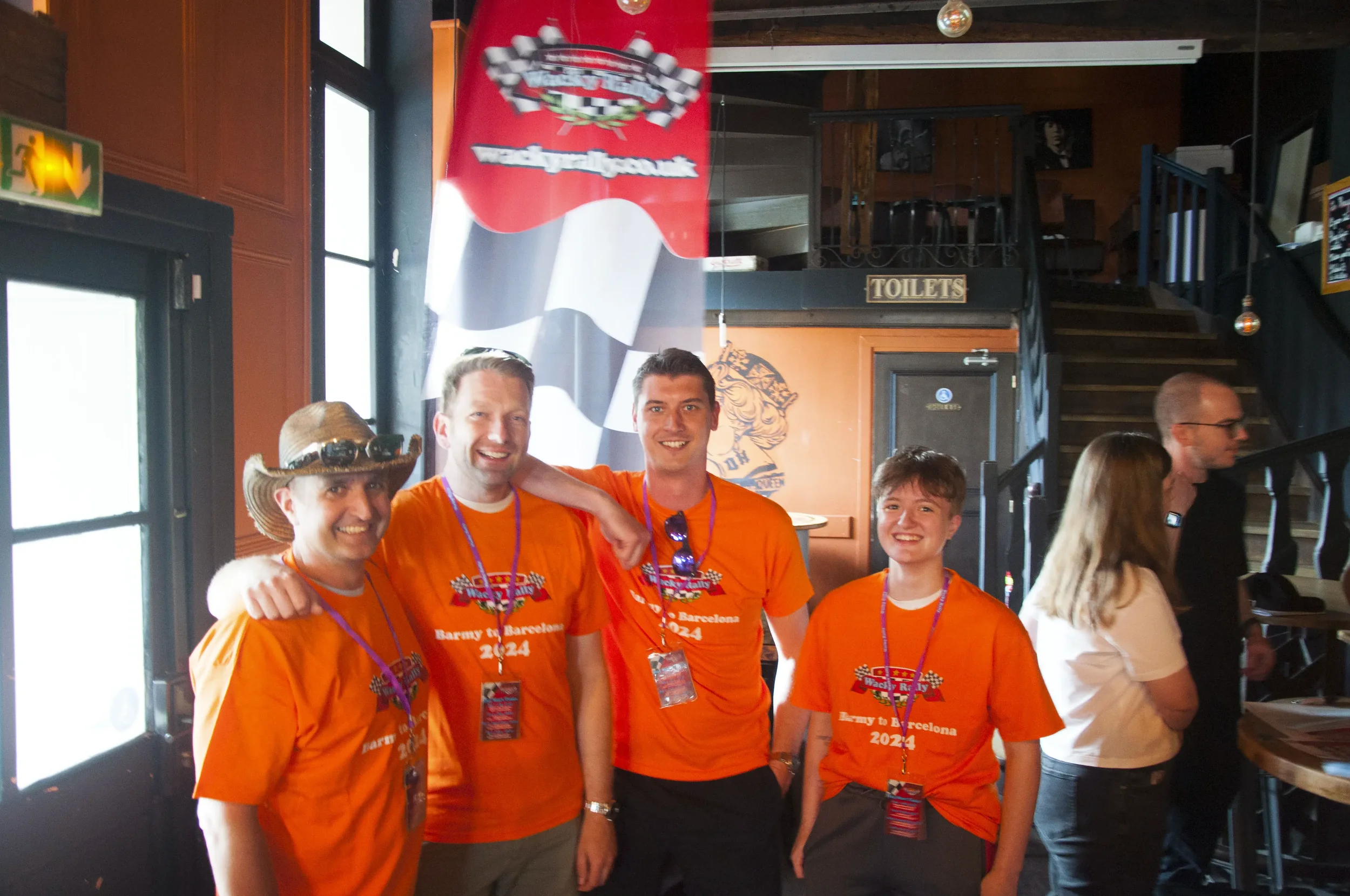 Four men and one woman at an indoor event wearing orange t-shirts with the text 'Barcy Barcelona 2024' and racing-themed graphics. The men are smiling, and the woman is smiling as well, with the man on the far left wearing a cowboy hat and goggles. T