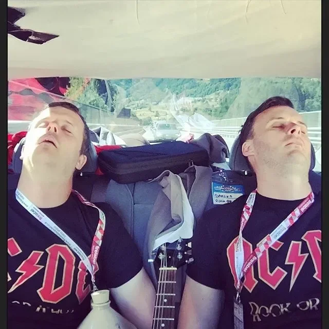 Two men wearing AC/DC shirts sleeping in the backseat of a car, with backpacks and a guitar between them.