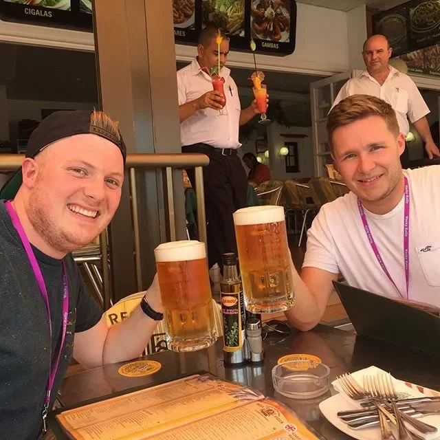 Two young men sitting at a restaurant table, each holding a large glass of beer, smiling at the camera. Two waiters in the background holding colorful cocktails, inside a casual dining setting with menus, condiments, and utensils on the table.