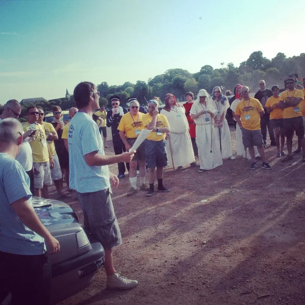 Group of people gathered outdoors, listening to a person reading from a paper. Many individuals are wearing yellow shirts, and some are dressed in white robes. The setting appears to be a sports field or open area with a natural background and a sunn