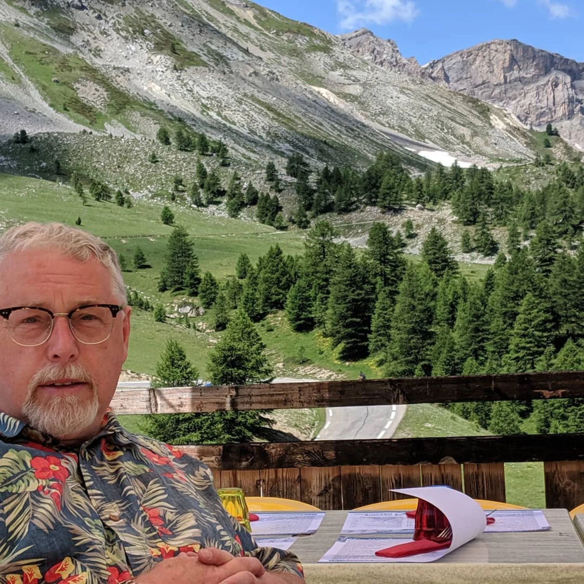 Man with glasses and a beard sitting at an outdoor table on a wooden deck with a mountainous landscape in the background, including green trees, rocky slopes, and a winding road.