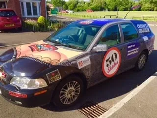A car with a promotional wrap advertising 50% discounts, parked on a street in a suburban area with houses and trees nearby.