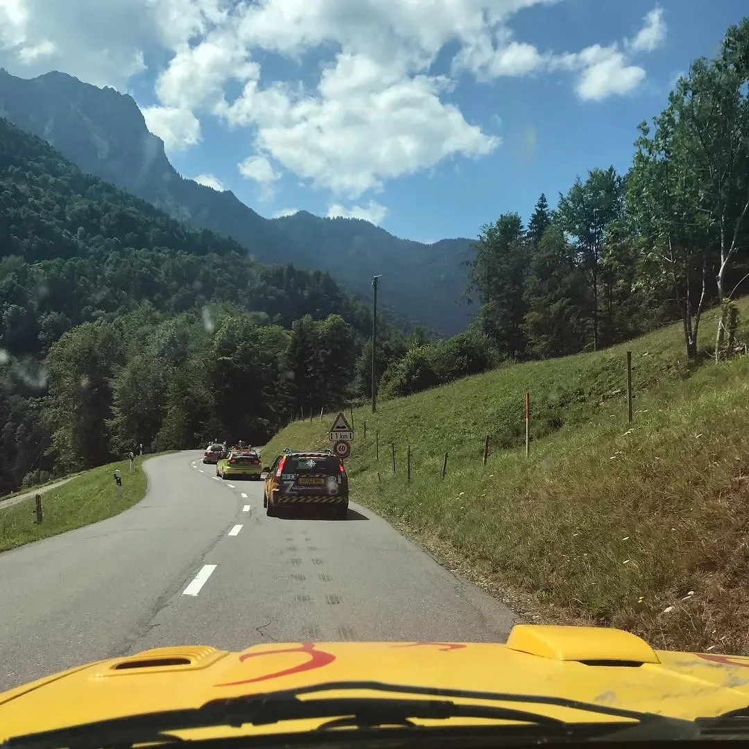 A winding mountain road with several cars parked along the side, surrounded by green trees and grass, with mountains and partly cloudy sky in the background.