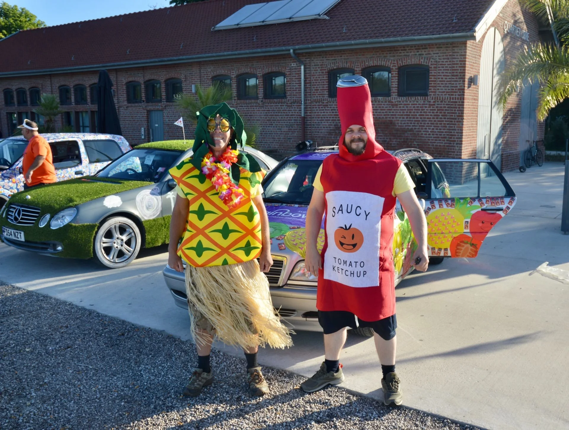 Two people dressed in food-themed costumes standing in front of decorated cars at a car show or parade. One is dressed as a pineapple with a grass skirt, and the other as a bottle of ketchup.