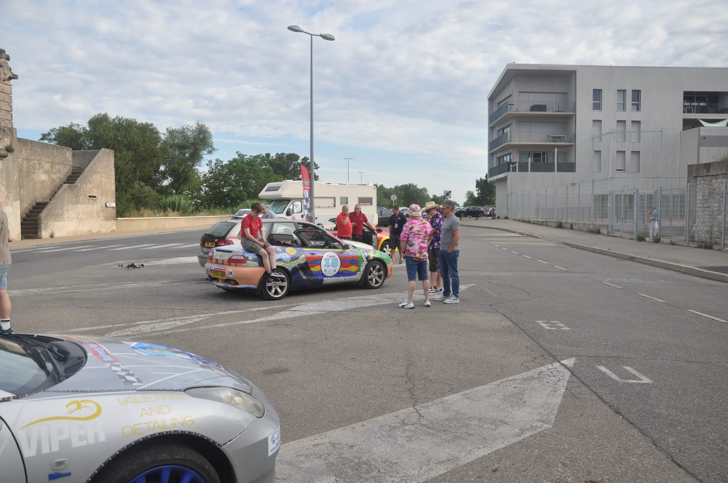 People gathered around a colorful race car in a parking lot with a camper van and a modern building in the background.