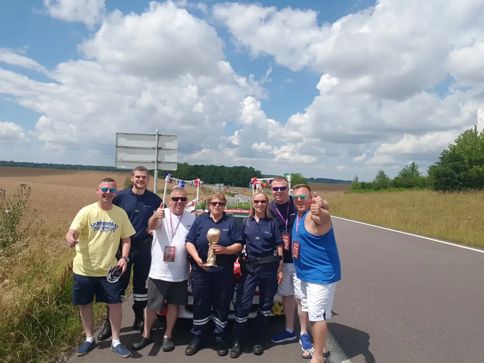 Group of people celebrating on the side of a highway, some wearing police uniforms, holding a trophy, with a decorated vehicle in the background. Clear sky with scattered clouds.