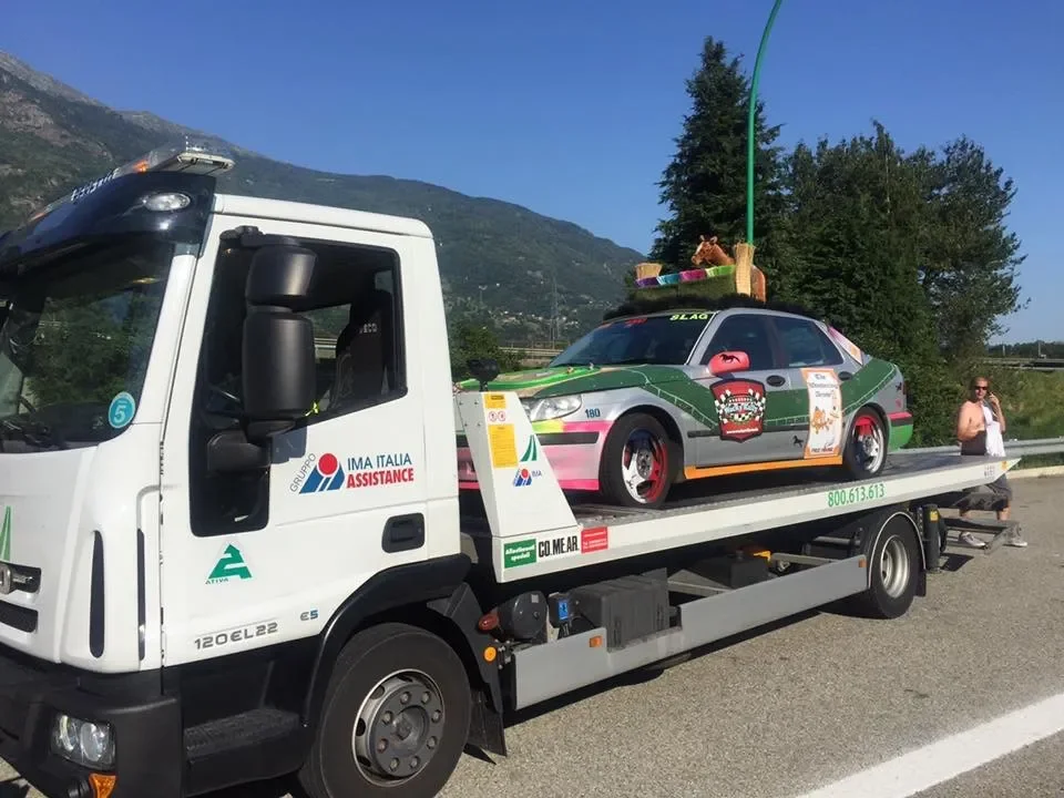 A tow truck carrying a race car decorated like a festive float, with a person standing nearby on the side of the road.